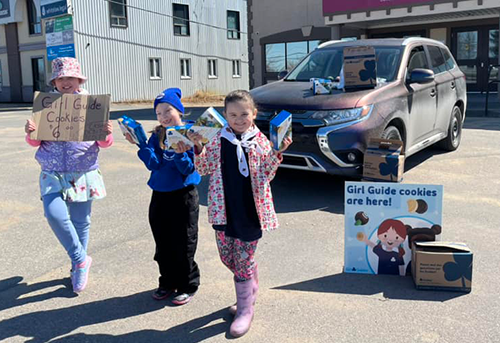 Three girls in a parking lot with signs selling cookies.