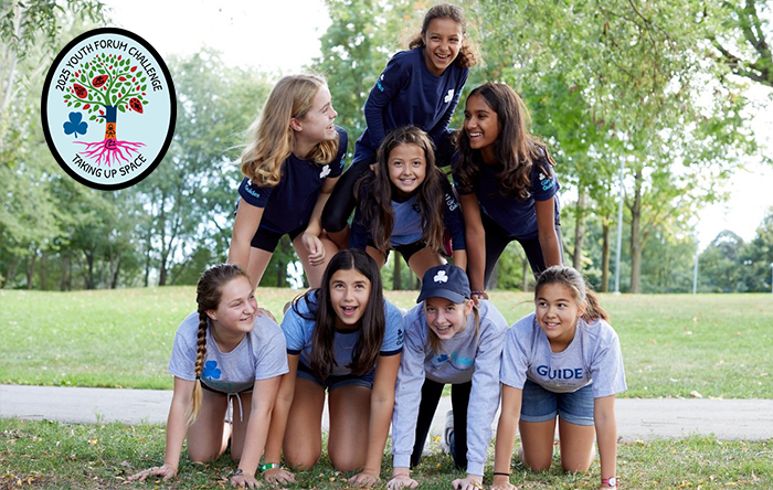 Eight girls laughing in a field forming a human pyramid with the Taking Up Space crest overlayed on the top left corner of the image.