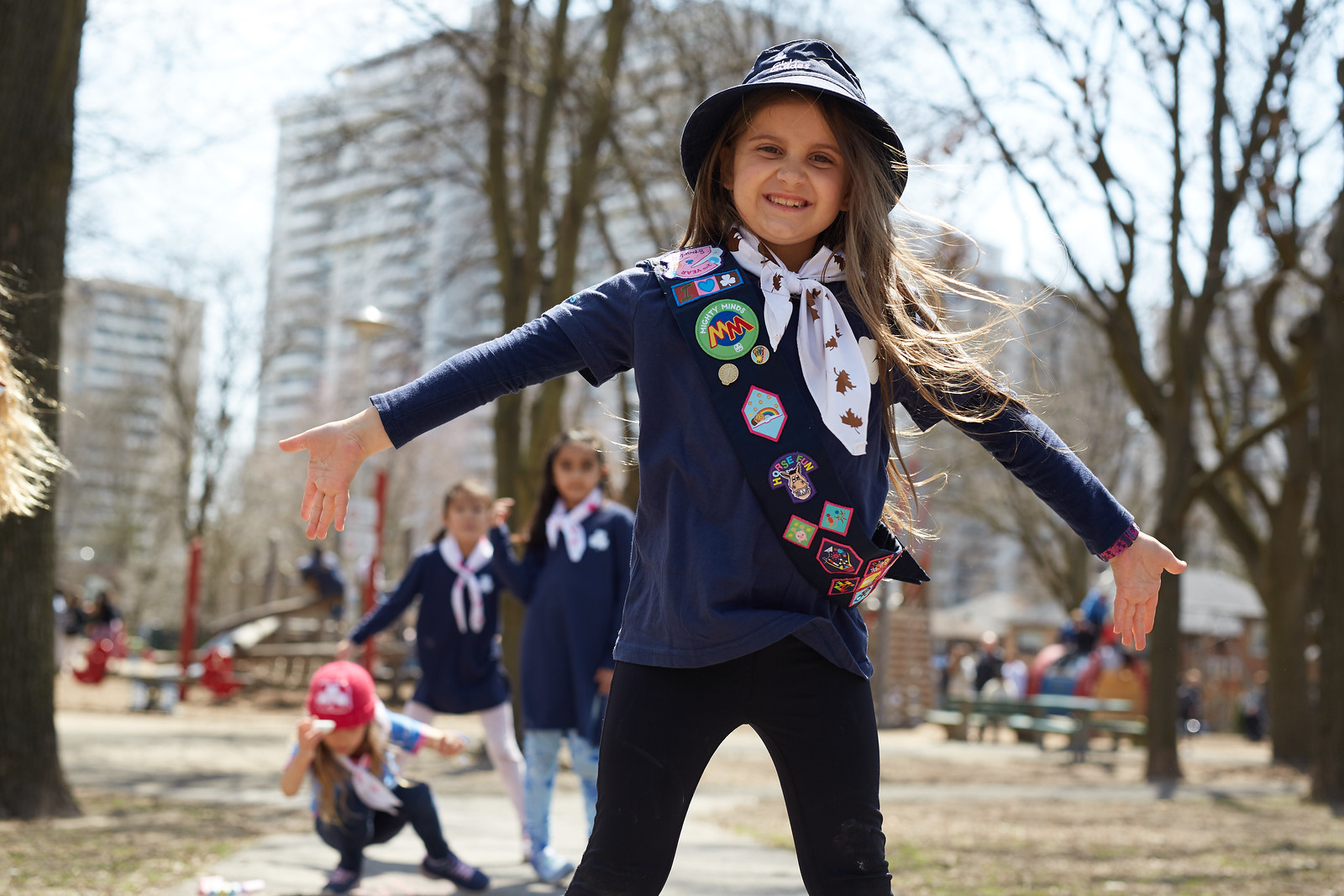 A Girl Guide aged 7 to 8 smiling and posing with arms outsretched. She is outside at a park with friends playing behind her