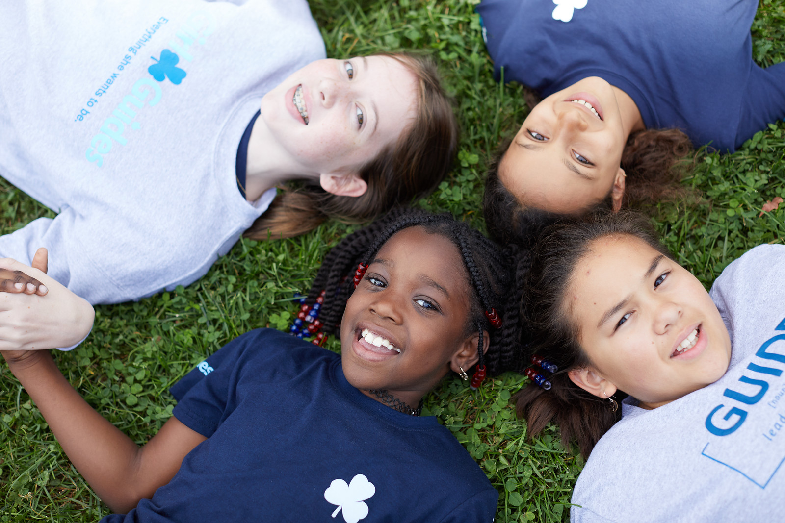 Four Girl Guides aged 9 to 11 lying in a circle in the grass outside, holding hands and smiling