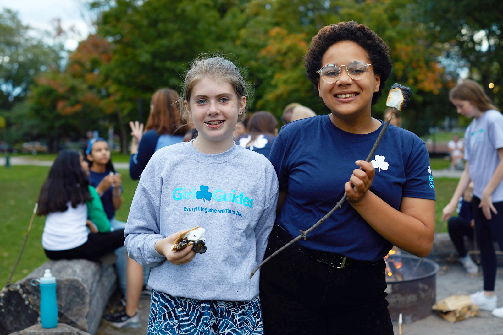 Two Girl Guides age 12 to 14 standing outside near a campfire holding s’mores and roast marshmallows on a stick